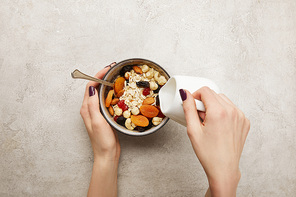 cropped view of woman pouring milk from milk jug into bowl with muesli, dried apricots and berries, nuts on textured grey surface