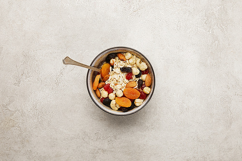 top view of bowl with muesli, dried apricots and berries, nuts and spoon on textured grey surface