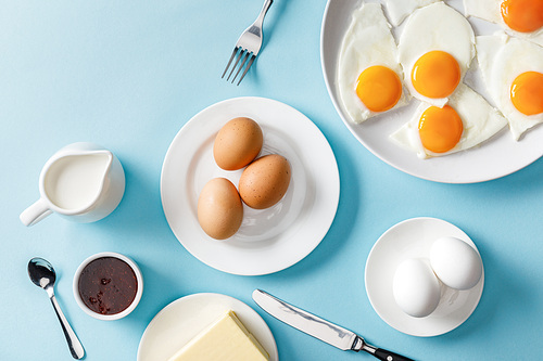 top view of boiled and fried eggs, butter, jam on white plates, milk, fork, spoon and knife on blue background