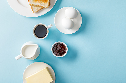top view of fresh eggs, jam, butter, coffee, milk and two toasts on white plates on blue background