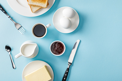 top view of fresh eggs, jam, coffee, cutlery, butter and two toasts on white plates on blue background