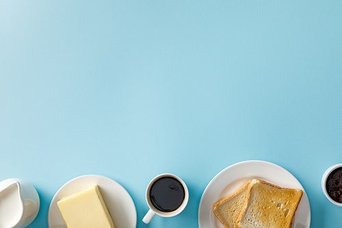 top view of milk, butter, jam, cup of coffee and two toasts on white plates on blue background