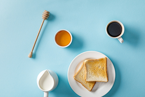 top view of honey in bowl, wooden dipper, coffee, milk and two toasts on white plate on blue background