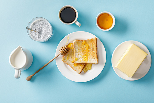 top view of coffee, milk, yogurt with chia seeds, butter, honey, toasts with honey and wooden dipper on white plates on blue background
