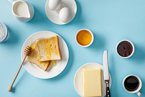 top view of served breakfast with yogurt, milk, coffee, jam, honey, butter and knife, toasts on white plates on blue background