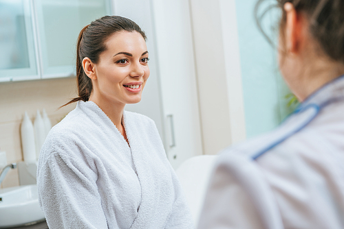 Beautiful smiling young woman in white bathrobe looking at cosmetologist in beauty clinic