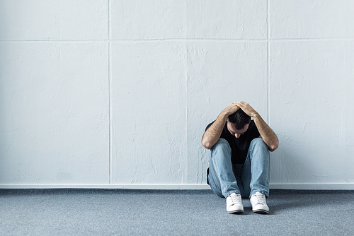 depressed man sitting on floor near white wall and holding hands on head