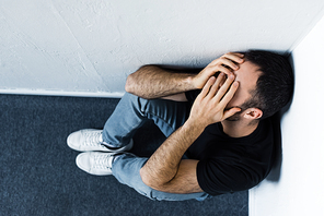 top view of upset man sitting on floor and corner and covering face with hands