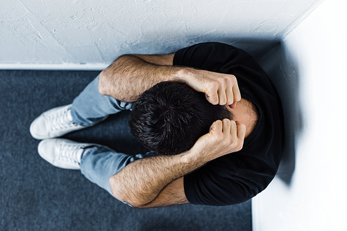 top view of adult depressed man sitting on floor in corner and holding hands on head