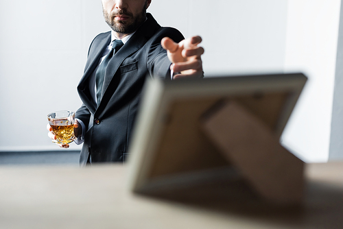 selective focus of man in black suit holding glass of whiskey and outstretching hand to desk with photo frame