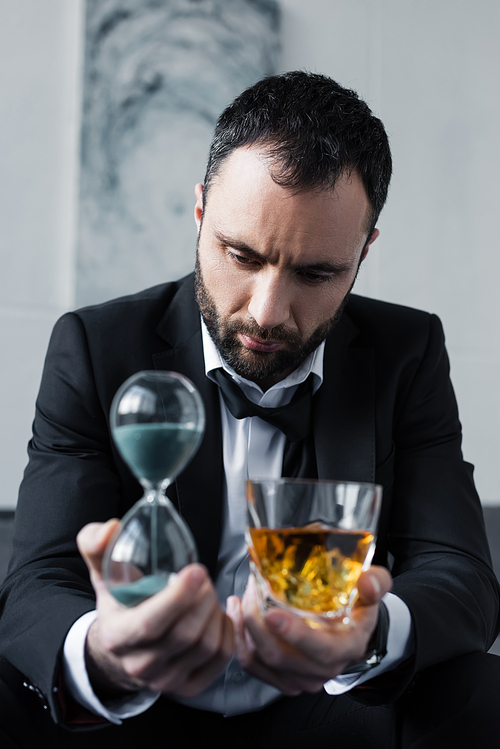 selective focus of depressed businessman holding glass of whiskey and hourglass