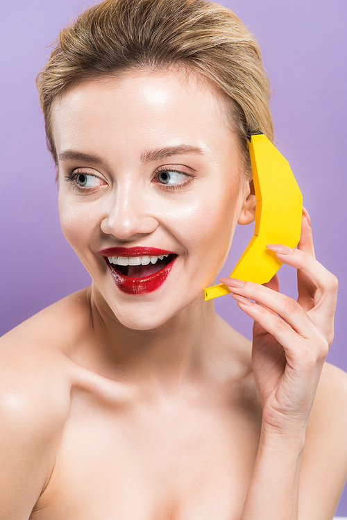 happy naked young woman holding yellow decorative banana near ear isolated on purple