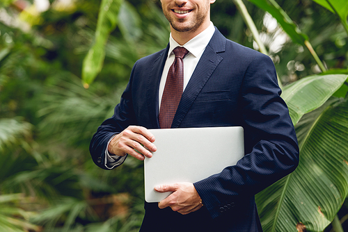 cropped view of smiling businessman in suit and tie holding laptop in orangery