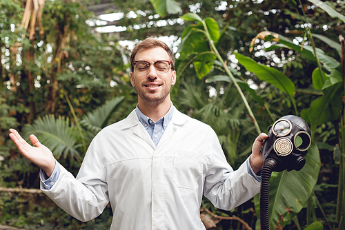 smiling handsome scientist in white coat and glasses with closed eyes holding rubber gas mask in orangery