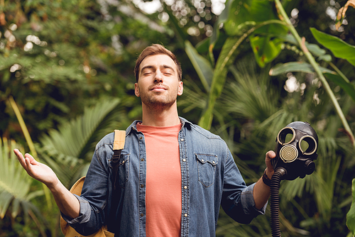 smiling traveler with backpack and closed eyes breathing fresh air while holding gas mask in tropical forest