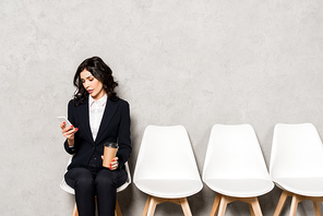 attractive woman using smartphone while holding paper cup and sitting on chair