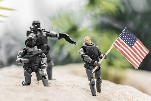 selective focus of toy soldiers aiming with guns at toy man with american flag on sand hill