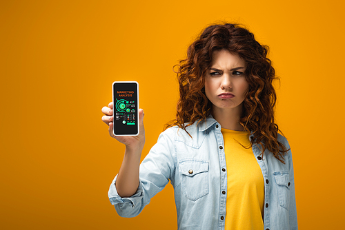 upset redhead woman holding smartphone with marketing analysis on screen on orange