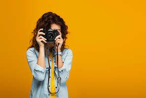 curly redhead photographer covering face with digital camera isolated on orange