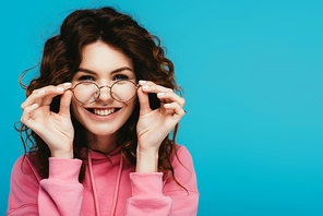 cheerful curly redhead girl touching glasses isolated on blue
