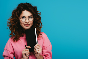 cheerful curly girl in glasses holding notebook and pen isolated on blue