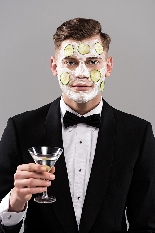 young man in formal wear with cucumber facial mask holding glass of cocktail isolated on grey