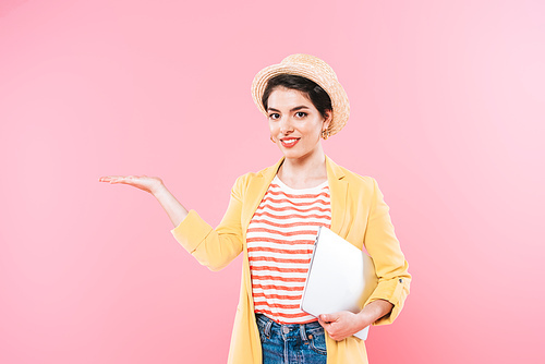 attractive mixed race woman holding laptop and gesturing isolated on pink