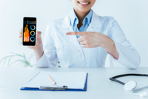 partial view of smiling latin doctor pointing with finger at smartphone with infographics on screen
