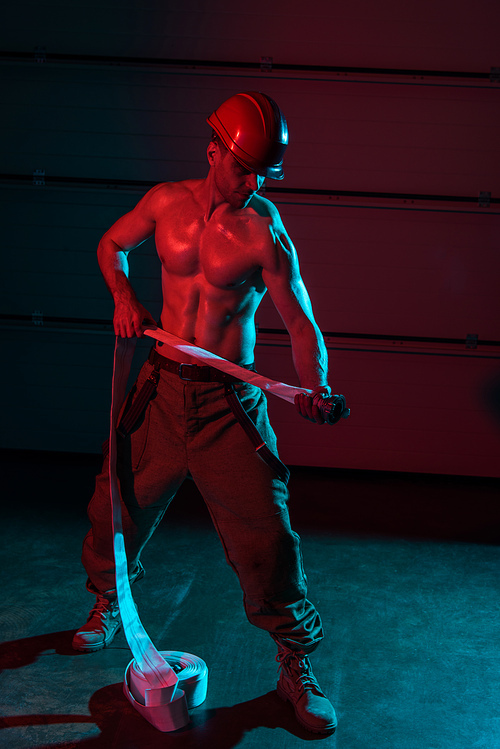 full length view of shirtless sexy fireman holding fire hose in darkness