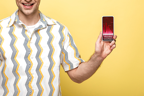 cropped view of smiling man showing smartphone with trading courses app, isolated on yellow