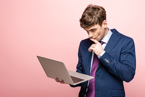thoughtful businessman in suit using laptop and holding hand near face on pink background
