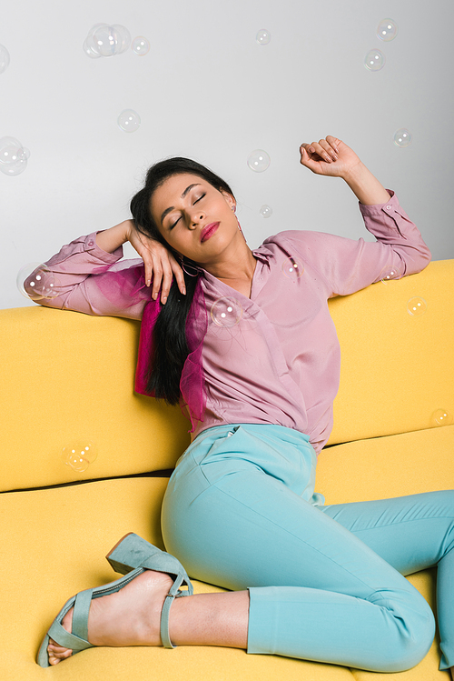 attractive brunette woman with closed eyes sitting on sofa near soap bubbles on white