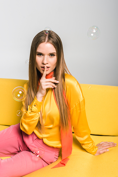 attractive blonde woman sitting on yellow sofa and showing hush sign on white