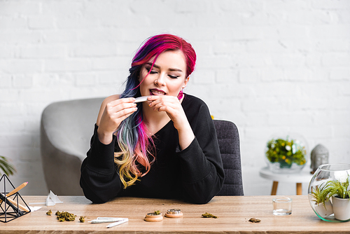 attractive girl licking joint with medical marijuana while sitting behind table in living room
