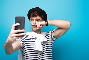 cheerful man taking selfie with smartphone while holding finger with drawn mustache near face on blue background