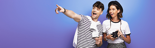 panoramic shot of cheerful man looking away and pointing with finger near smiling girl with digital camera on blue background