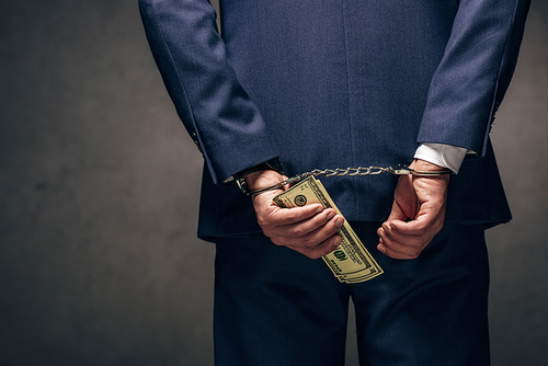 cropped view of handcuffed businessman in suit holding dollar banknotes on grey