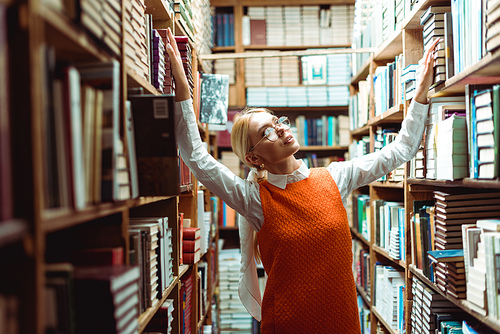 pretty and blonde woman in glasses and orange dress looking away in library