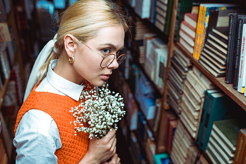 beautiful and blonde woman holding white flowers and looking away in library