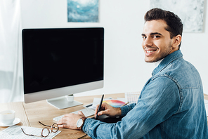 Designer smiling at camera while working on project of user experience design near computer and graphics tablet on table