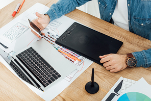 Cropped view of ux designer using smartphone near laptop, graphics tablet and sketches on table