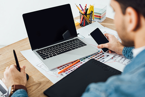 Selective focus of ux designer using smartphone near laptop, graphics tablet and layouts with sketches on table on grey background