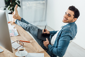 Barefoot ux designer smiling at camera while using graphics tablet and computer near color palette on table in office