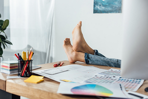 Cropped view of barefoot designer sitting at table with computer and layouts of user experience design