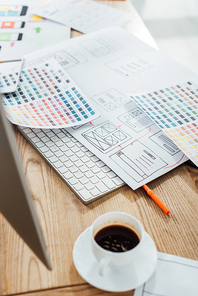 Selective focus of coffee cup and computer monitor near color palettes and sketches of ux design on wooden table