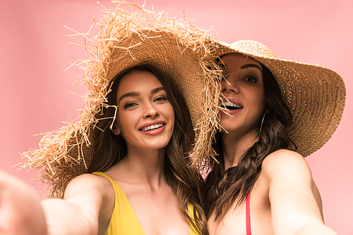 two smiling girls in straw hats taking selfie isolated on pink