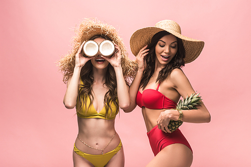 two smiling girls in straw hats holding coconuts and pineapple isolated on pink