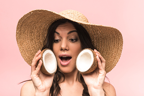 surprised woman in straw hat and swimsuit holding coconuts isolated on pink