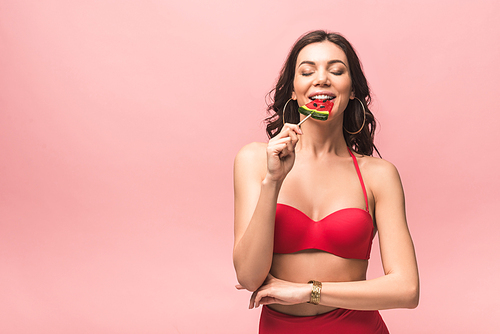 attractive young woman in swimsuit eating watermelon lollipop isolated on pink