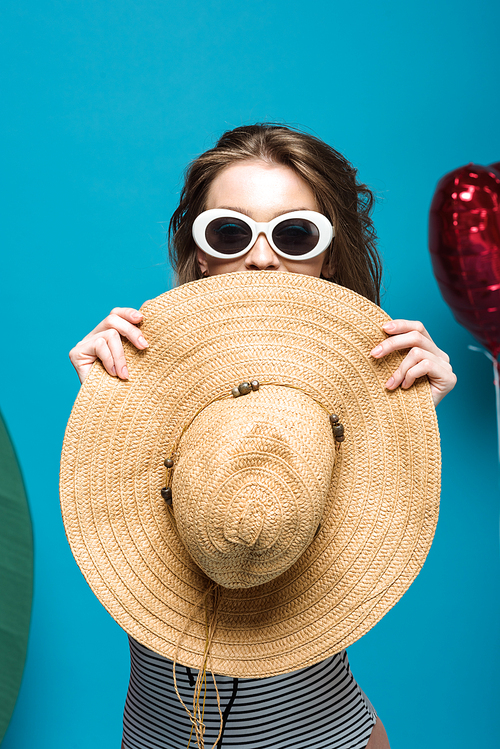 happy young woman in sunglasses holding straw hat on blue